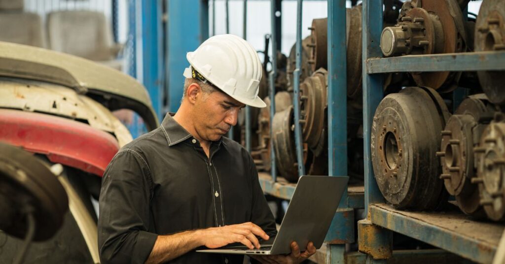 Car wrecker using a laptop while inspecting recycled vehicle parts stored at an auto recycling facility.